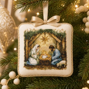 May include: A square-shaped fabric Christmas ornament featuring a cross-stitched nativity scene. The design shows figures in blue and white robes surrounding a central manger under a star, finished with a gold braided trim and a satin ribbon.