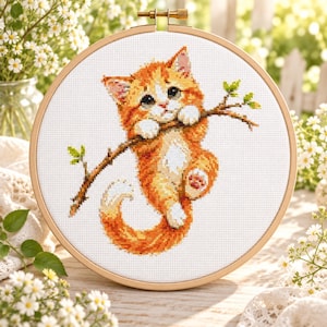 Puede incluir: Un bastidor de bordado circular de madera que contiene un patrón de punto de cruz de un gatito atigrado naranja colgado de una rama de árbol marrón con pequeñas hojas verdes. El gatito tiene patas blancas, pecho blanco y una cola rizada sobre un fondo de tela blanca.