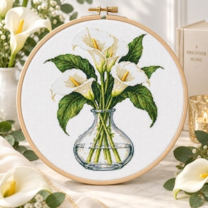 May include: A cross-stitch embroidery project in a round wooden hoop featuring a bouquet of white calla lilies with yellow spadices and green leaves arranged in a clear glass vase, displayed against a white fabric background.