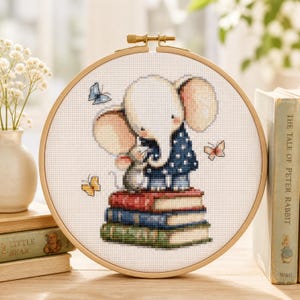 May include: A circular wooden embroidery hoop containing a cross-stitch pattern of a white elephant and a small gray mouse standing on a stack of three books. The elephant wears a dark blue polka-dot shirt. Three butterflies in blue, pink, and yellow surround them.