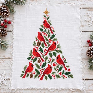May include: A white fabric piece featuring a cross-stitch design of a Christmas tree formed by green holly leaves, red berries, and several bright red cardinal birds. A gold star sits at the top of the tree, which is set on a white background.