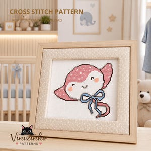 May include: A cross stitch pattern featuring a pink stingray with white polka dots, a smiling face, and a blue bow tied around its tail. The design is displayed in a light wood frame with a beige fabric border. Text reads CROSS STITCH PATTERN.