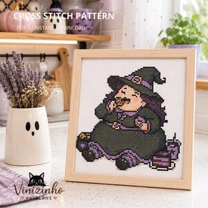 Op de afbeelding: Een kruissteekpatroon in een lichthouten lijst met een cartoonheks die een zwart-paarse hoed en jurk draagt en een koekje eet. Het ontwerp is geborduurd op witte Aida-stof. De tekst CROSS STITCH PATTERN PDF INSTANT DOWNLOAD is zichtbaar.