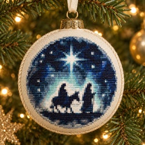 Puede incluir: Un adorno navideño redondo de tela color crema con una escena en punto de cruz que muestra la silueta de una persona montada en un burro y otra caminando bajo una estrella brillante y luminosa sobre un cielo nocturno azul estrellado.