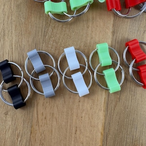 May include: A collection of small, rectangular plastic connectors in black, grey, white, light green, and red, each featuring a central indentation and attached to two circular metal split rings, arranged in pairs on a wooden surface.