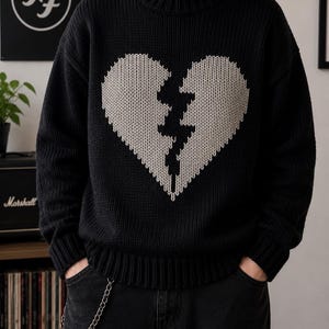 May include: A black long-sleeve knit sweater featuring a large, centered, light gray broken heart graphic on the chest. The sweater has a ribbed crew neckline, ribbed cuffs, and a ribbed hem, constructed with a thick, textured yarn weave.