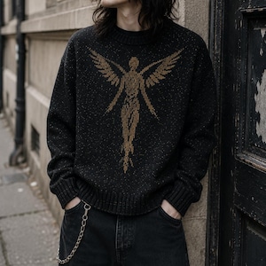 May include: A black speckled knit jumper with a gold angel design on the front. The jumper has a crew neck and long sleeves. The person in the image is wearing black jeans and a silver chain.