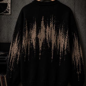 May include: A black long-sleeve knit sweater featuring a tan, jagged, vertical soundwave-like pattern across the chest and sleeves. The garment has a ribbed crew neckline, ribbed cuffs, and a ribbed hem, hanging on a wooden hanger.
