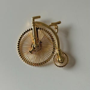 Op de afbeelding: Goudkleurige fietsbroche met een groot voorwiel en een kleiner achterwiel. De broche heeft ingewikkelde spaakdetails en een klassiek hoge-fietsontwerp, perfect om een vleugje vintage charme toe te voegen.