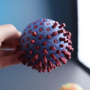 Op de afbeelding: Een bolvormig object met een matblauw oppervlak voorzien van een raster van kleine, vierkante rode uitsteeksels die naar buiten steken. Het object is opgebouwd uit meerdere in elkaar grijpende segmenten met zichtbare 3D-printlijnen.