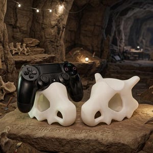 Op de afbeelding: Een zwarte videogamecontroller rustend op een witte, holle, botvormige standaard, vergezeld door een tweede identieke witte standaard. De standaards hebben een gestileerd, prehistorisch schedelontwerp met holle oogkassen en neusholtes.