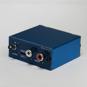 Könnte beinhalten: Eine blaue PHONO-EQ-100 Vorverstärkerbox mit einem Kippschalter, einem USB-Typ-C-Ladeanschluss und zwei RCA-Ausgangsbuchsen, die mit L und R gekennzeichnet sind. Das Gerät hat ein kompaktes, rechteckiges Metallgehäuse mit weißem Aufdruck.