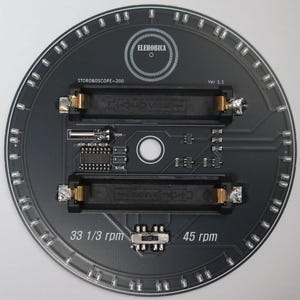 May include: A circular black circuit board with the text "ELEHOBICA" and "STOROBOSCOPE-200". It features two rectangular slots, various electronic components, and the text "33 1/3 rpm" and "45 rpm". A ring of small lights surrounds the edge.