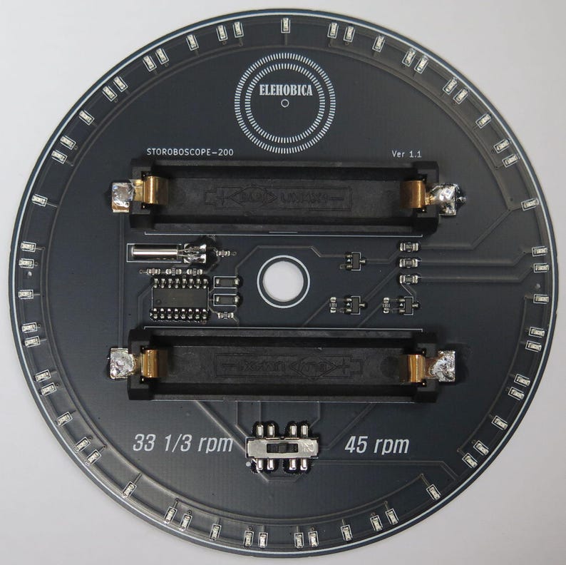 May include: A circular black circuit board with the text "ELEHOBICA" and "STOROBOSCOPE-200". It features two rectangular slots, various electronic components, and the text "33 1/3 rpm" and "45 rpm". A ring of small lights surrounds the edge.