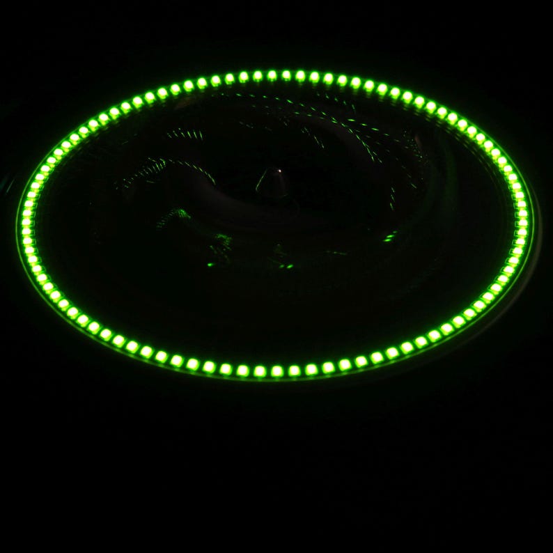 May include: A circular LED light ring glows bright green against a black background. The ring is composed of numerous small, evenly spaced LED lights, creating a continuous illuminated effect. The overall design is modern and sleek.