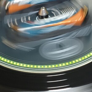 May include: Close-up of a rotating object with a silver central component. The object has a dark gray surface with orange and blue accents. A ring of green lights encircles the base, creating a dynamic and modern aesthetic.