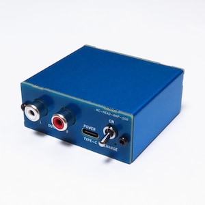 Könnte beinhalten: Ein blauer, rechteckiger Audioverstärker mit der Aufschrift MC-HEAD-AMP-100, ausgestattet mit zwei Cinch-Ausgangsbuchsen, einem USB-Typ-C-Stromanschluss und einem Metallkippschalter. Das Gerät hat ein blaues Gehäuse mit weißer Beschriftung.