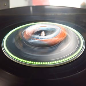 May include: A close-up shot of a black turntable with a spinning record. The record's center features a silver spindle and a blurred, colorful design of orange, blue, and white. A ring of green lights encircles the record's edge.
