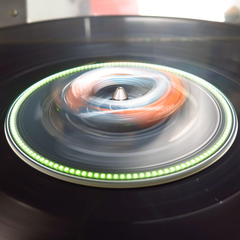 May include: A close-up shot of a black turntable with a spinning record. The record's center features a silver spindle and a blurred, colorful design of orange, blue, and white. A ring of green lights encircles the record's edge.