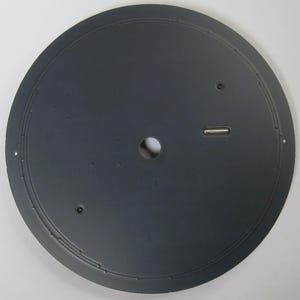 May include: A circular, dark gray metal object with a central hole and a small, rectangular metal piece. The object has a series of small holes around the edge and a circular groove near the edge. The surface appears to be matte.