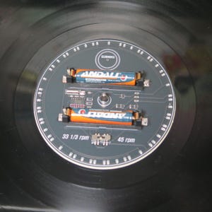 May include: A black vinyl record with a gray circuit board design in the center. Two orange and blue batteries are mounted on the board. The text "ELEHORICA", "33 1/3 rpm", and "45 rpm" are visible.