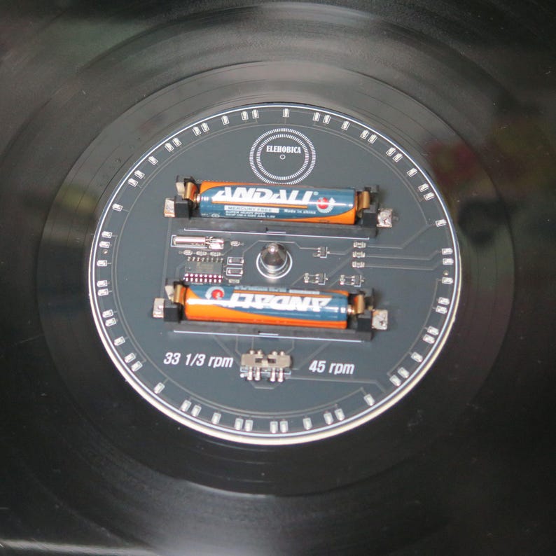 May include: A black vinyl record with a gray circuit board design in the center. Two orange and blue batteries are mounted on the board. The text "ELEHORICA", "33 1/3 rpm", and "45 rpm" are visible.