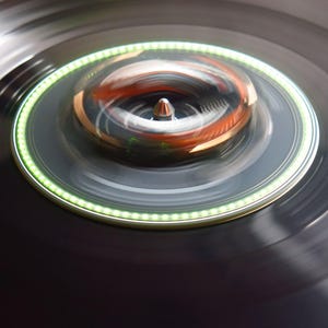 May include: Close-up of a spinning record player with a black vinyl record. The turntable features a green illuminated ring and a silver spindle. The center of the record player has a copper-colored element. The image has a blurred effect, suggesting motion.