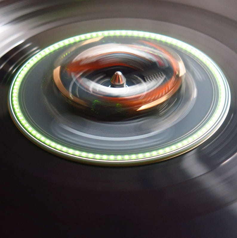May include: Close-up of a spinning record player with a black vinyl record. The turntable features a green illuminated ring and a silver spindle. The center of the record player has a copper-colored element. The image has a blurred effect, suggesting motion.