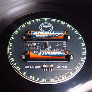 May include: A black circular device with a circuit board design, featuring two orange and blue batteries. The device has a ring of green lights and the text "ELENOBICA". It also displays "33 1/3 rpm" and "45 rpm".