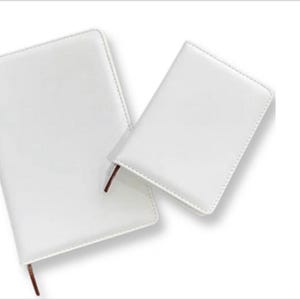 May include: Two white leather journals are shown, one larger than the other. Both have a smooth, glossy finish and a brown ribbon bookmark. The journals are angled against a white background.