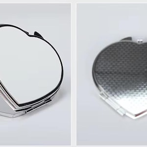 May include: Heart-shaped compact mirror with a silver-tone metal frame. The mirror is open, revealing the reflective surface. The back of the compact has a textured pattern. The image also shows the text "M6".