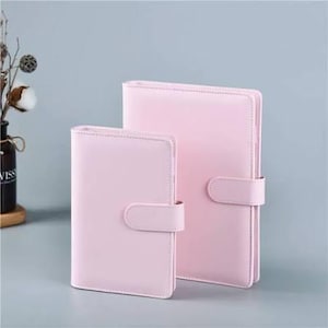 May include: Two light pink faux leather journals with rounded corners and a tab closure. The journals are different sizes and are standing upright against a light gray background. The journals are a solid color.