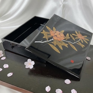 以下が含まれることがあります： 光沢のある黒い漆塗りの長方形の箱。蓋には、細長い葉と小さな花の房が描かれた、金と銅の色調の花の枝のデザインが施されています。右下隅には小さな赤い四角い印が見えます。
