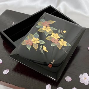 May include: A rectangular black lacquer box with a glossy finish, featuring a painted floral design of yellow and muted red blossoms with green leaves. The lid includes a small red seal and Japanese characters in the bottom right corner.