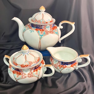 Puede incluir: Un juego de té de porcelana blanca con tetera, azucarero y jarrita para la leche. El juego presenta un diseño floral con detalles en rojo, azul y dorado. La tetera tiene un pico y asa curvados.