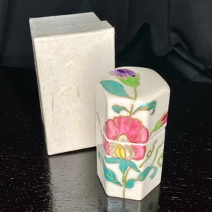 Op de afbeelding: Een zeshoekige witte keramische pot met deksel, voorzien van een handgeschilderd bloemmotief met een grote roze bloem, groene bladeren en een paarse knop. De pot wordt geleverd met een eenvoudige, rechthoekige witte kartonnen doos.