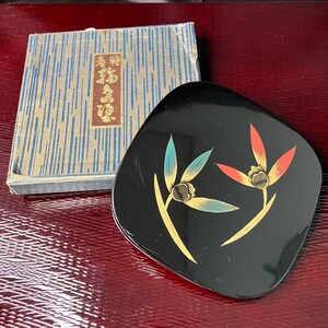 Puede incluir: Un plato cuadrado de laca negra con esquinas redondeadas que presenta un diseño pintado de dos flores estilizadas en tonos azul verdoso, naranja y dorado. Junto al plato hay una caja con estampado azul y dorado con texto japonés en una etiqueta roja.