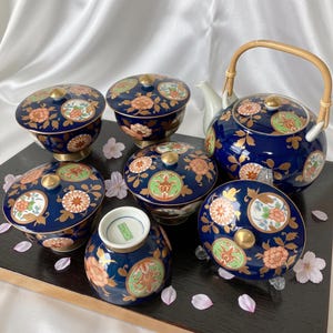 May include: A tea set featuring a dark blue teapot with a bamboo handle and four matching lidded bowls. Each piece is decorated with gold floral patterns and circular medallions containing orange, green, and white chrysanthemum and peony motifs.