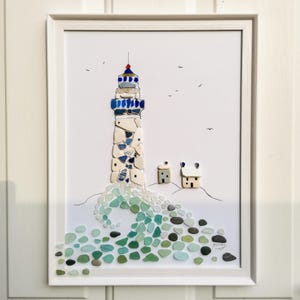 Op de afbeelding: Een ingelijst kunstwerk aan de muur met een vuurtoren en twee kleine huisjes gemaakt van zeeglas en kiezels. De vuurtoren heeft blauwe en witte segmenten, terwijl de basis een brekende golf van groen, blauw en grijs zeeglas toont.
