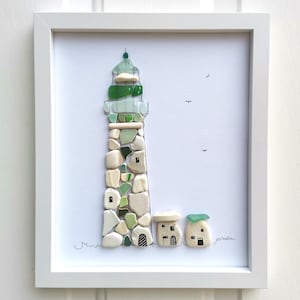 May include: A framed wall art piece featuring a lighthouse and two small buildings constructed from white, cream, and green sea glass and pebbles. The design includes hand-drawn black ink details for windows, doors, and birds on a white background.