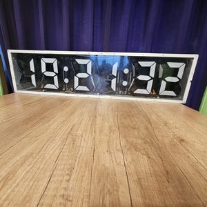 Op de afbeelding: Een rechthoekige digitale klok met een witte omlijsting en een zwart displaypaneel met grote, witte, gesegmenteerde cijfers die de tijd 19:21:32 weergeven. De klok heeft een doorzichtige voorkant en zichtbare interne elektronische onderdelen.