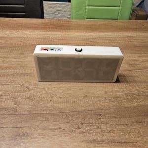 Puede incluir: Un reloj digital rectangular de color blanco con acabado liso. La superficie superior presenta un pequeño botón circular negro y tres puertos circulares empotrados de colores rojo, azul y blanco. El frontal es un panel translúcido con rejilla.