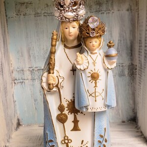 May include: A white porcelain figurine of the Virgin Mary and baby Jesus. Mary is wearing a blue and white robe with a gold crown and holding a scepter. Jesus is wearing a blue and white robe and holding a globe. The figurine is standing on a gold base with the inscription "Consolatrix Afflictorum".