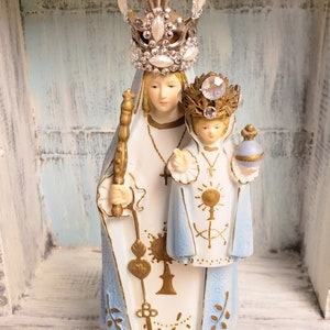 May include: A white porcelain figurine of the Virgin Mary and baby Jesus. Mary is wearing a blue robe with gold accents and a crown of rhinestones. Jesus is wearing a white robe with gold accents and a crown of rhinestones. The figurine is standing on a gold base.