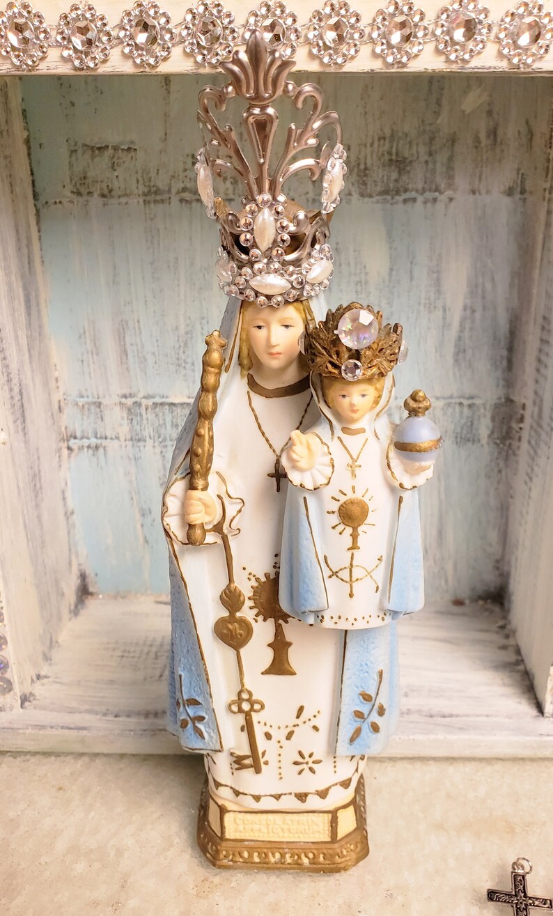 May include: A white porcelain figurine of the Virgin Mary and baby Jesus. Mary is wearing a blue robe with gold accents and a crown of rhinestones. Jesus is wearing a white robe with gold accents and a crown of rhinestones. The figurine is standing on a gold base.