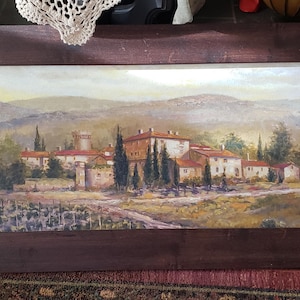 May include: A framed oil painting depicting a landscape scene with a village nestled in a valley. The painting features a cluster of buildings with red tile roofs, surrounded by trees and rolling hills. The sky is a soft blue with hints of white clouds.