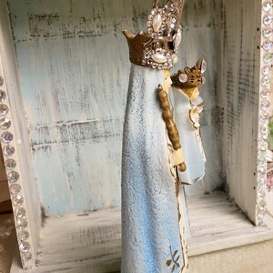 May include: A blue and gold figurine of the Virgin Mary and baby Jesus. The figurine is wearing a crown and a blue robe. The figurine is standing in a white wooden box with rhinestones around the edge.