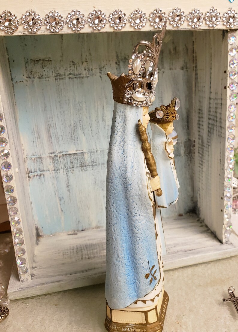 May include: A blue and gold figurine of the Virgin Mary and baby Jesus. The figurine is wearing a crown and a blue robe. The figurine is standing in a white wooden box with rhinestones around the edge.