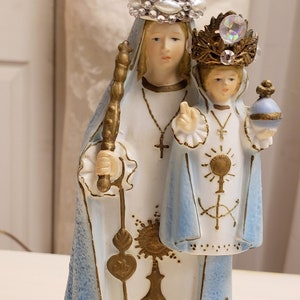May include: A white porcelain figurine of the Virgin Mary and baby Jesus. The Virgin Mary is wearing a blue robe and a silver crown with white gems. The baby Jesus is wearing a blue robe and a gold crown with a clear gem. The figurine is standing on a gold base with the inscription "Consolatrix Afflictorum".