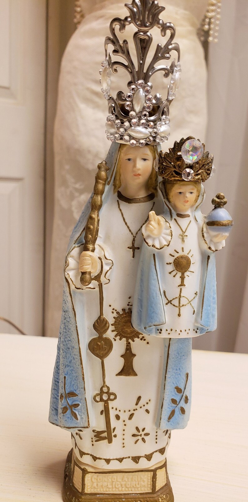 May include: A white porcelain figurine of the Virgin Mary and baby Jesus. The Virgin Mary is wearing a blue robe and a silver crown with white gems. The baby Jesus is wearing a blue robe and a gold crown with a clear gem. The figurine is standing on a gold base with the inscription "Consolatrix Afflictorum".